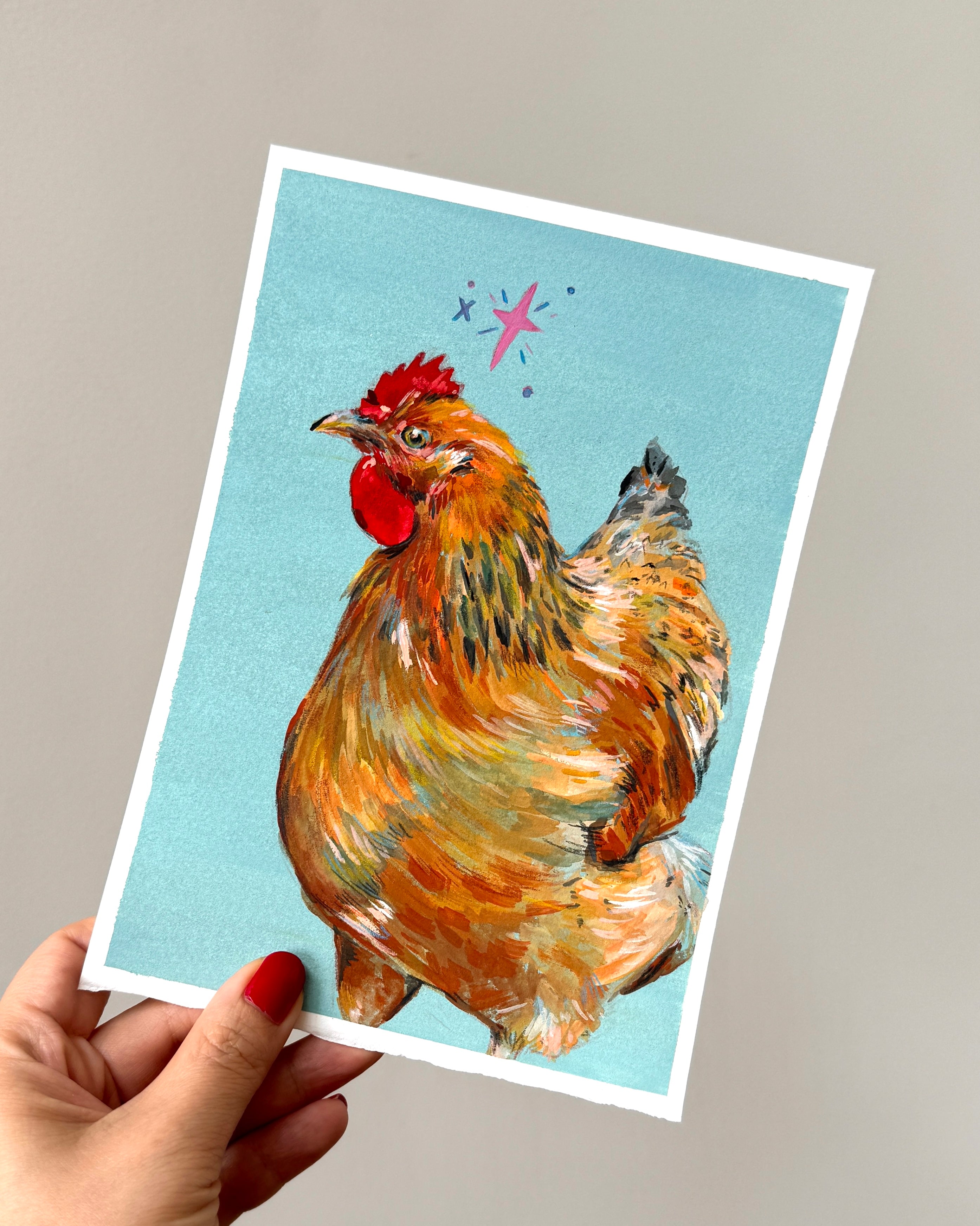 Hand holding a painting with a colourful illustration of a rooster on a soft teal background.