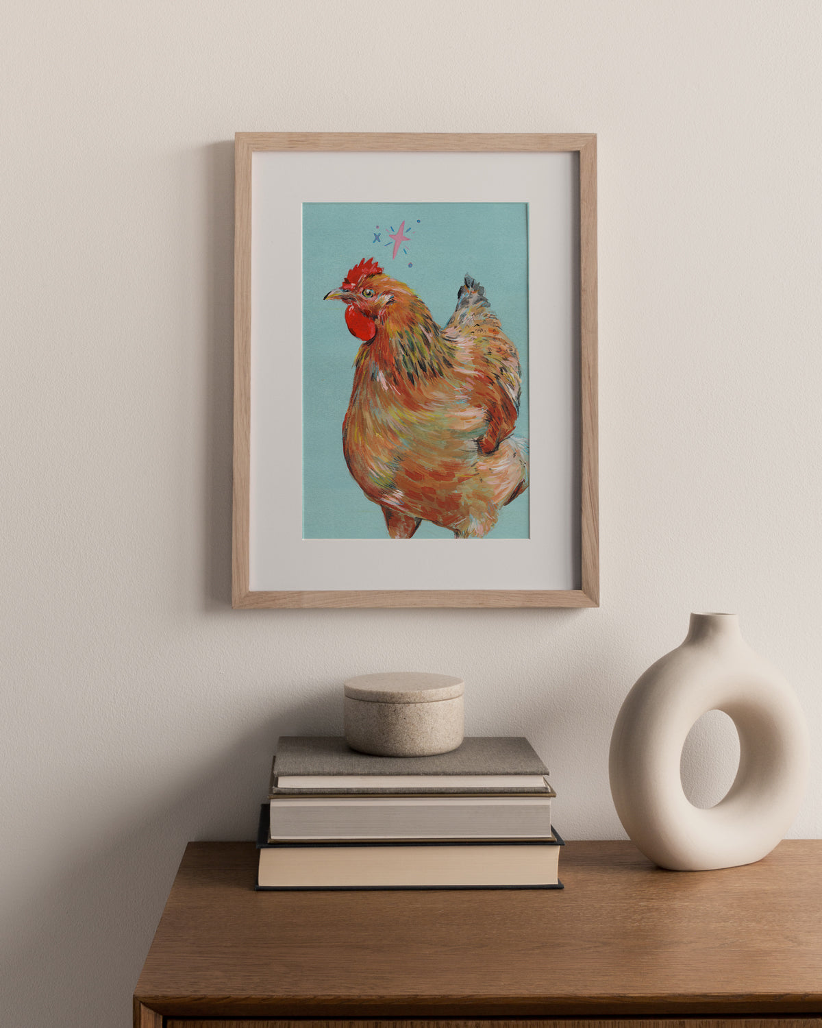 Framed artwork of a hen on a wall above books and a vase.
