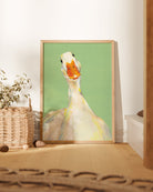 Framed artwork of a goose on a green background in a room with a basket and plant.